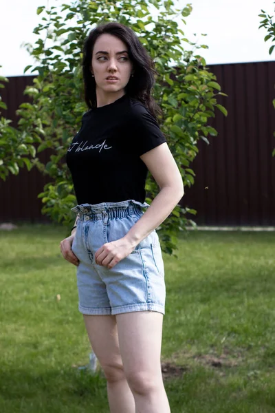 Encantadora jovem morena em uma camiseta preta e shorts jeans. No campo, no jardim . — Fotografia de Stock