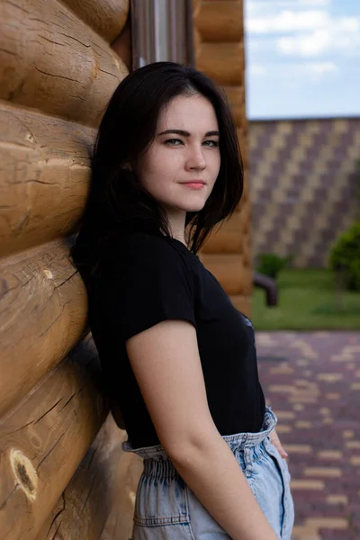 Aantrekkelijke jonge brunette vrouw in zwart t-shirt en spijkerbroek naast een houten modern huis — Stockfoto