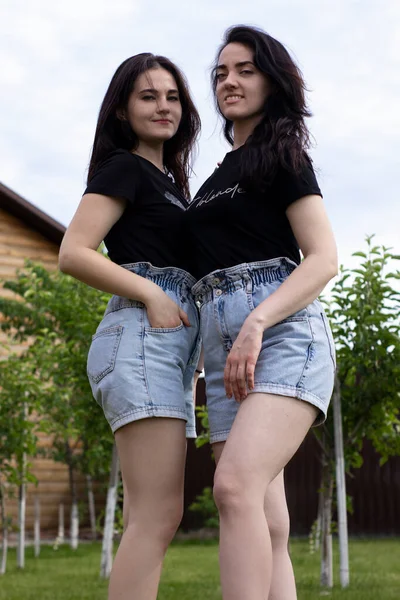 黒のTシャツとデニムのショートパンツに身を包んだ2人の若いブルネットの女性。姉妹・恋人・家族・友情の概念 — ストック写真
