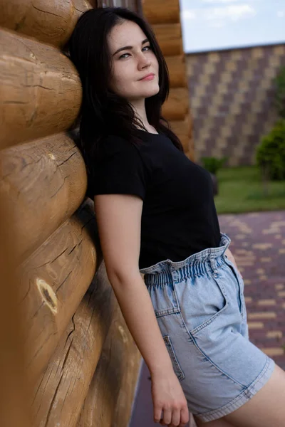 Atraente jovem morena mulher em t-shirt preta e calções jeans ao lado de uma casa moderna de madeira — Fotografia de Stock