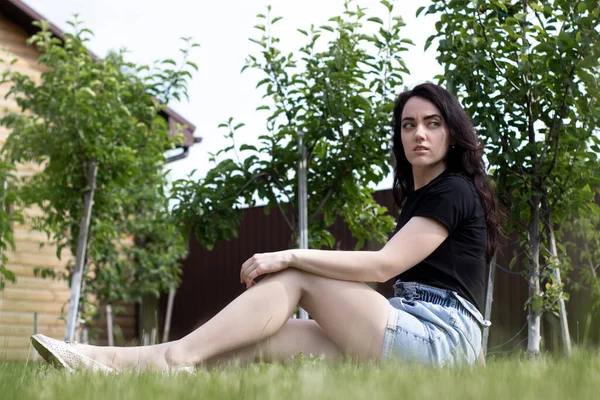 Encantadora jovem morena em uma camiseta preta e shorts jeans. No campo, no jardim . — Fotografia de Stock