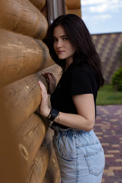 Atraente jovem morena mulher em t-shirt preta e calções jeans ao lado de uma casa moderna de madeira — Fotografia de Stock