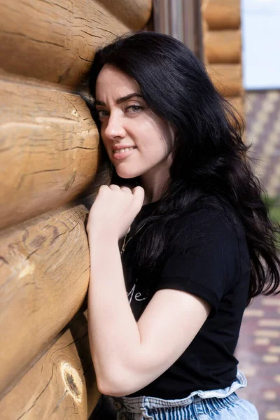 Aantrekkelijke jonge brunette vrouw in zwart t-shirt en spijkerbroek naast een houten modern huis — Stockfoto