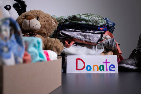 Donate sign handwritten with multicoloured letters. A box with clothes and a pile of clothes nearby on a grey table.clothes donation concept. copy space