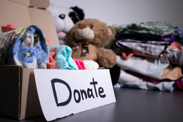 Donate sign handwritten with black letters. A box with clothes and a pile of clothes nearby on a grey table.clothes donation concept. copy space