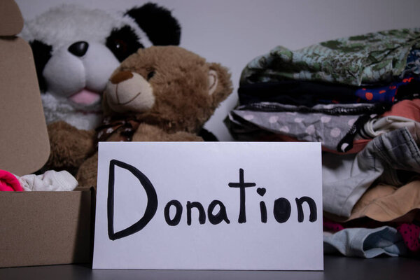 donation sign handwritten with black letters. A box with clothes and a pile of clothes nearby on a grey table.clothes donation concept. copy space