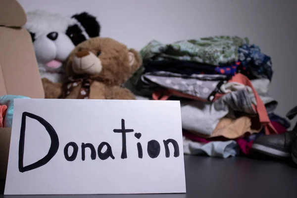 Spendenschild handgeschrieben mit schwarzen Buchstaben. Eine Schachtel mit Kleidung und ein Stapel Kleider in der Nähe auf einem grauen Tisch. Kopierraum — Stockfoto