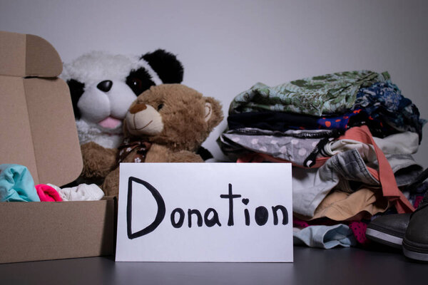 donation sign handwritten with black letters. A box with clothes and a pile of clothes nearby on a grey table.clothes donation concept. copy space