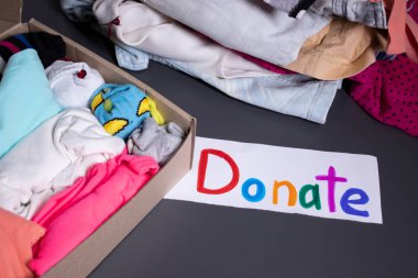 Çok renkli harflerle el yazısıyla yazılmış bir bağış yazısı. Gri bir masa. Kıyafet bağışı konsepti üzerine giysi ve giysi yığını. Boşluğu kopyala