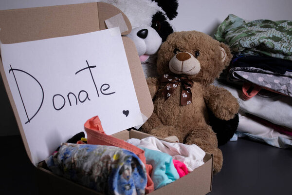 Donate sign handwritten with black letters. A box with clothes and a pile of clothes nearby on a grey table.clothes donation concept. copy space