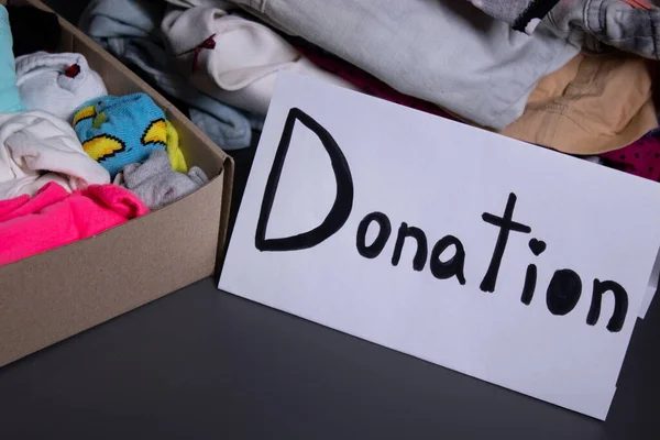 donation sign handwritten with black letters. A box with clothes and a pile of clothes nearby on a grey table.clothes donation concept. copy space