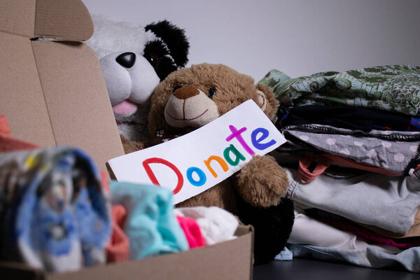 Donate sign handwritten with multicoloured letters. A box with clothes and a pile of clothes nearby on a grey table.clothes donation concept. copy space