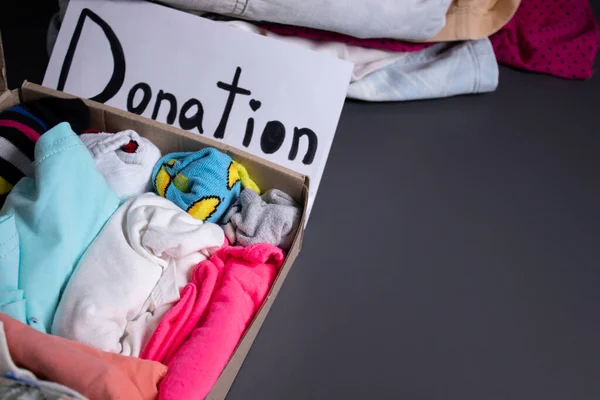 donation sign handwritten with black letters. A box with clothes and a pile of clothes nearby on a grey table.clothes donation concept. copy space