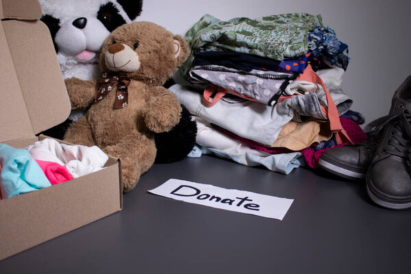 Donate sign handwritten with black letters. A box with clothes and a pile of clothes nearby on a grey table.clothes donation concept. copy space
