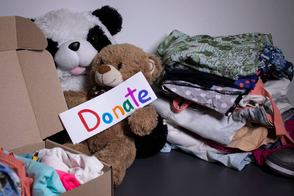 Donate sign handwritten with multicoloured letters. A box with clothes and a pile of clothes nearby on a grey table.clothes donation concept. copy space