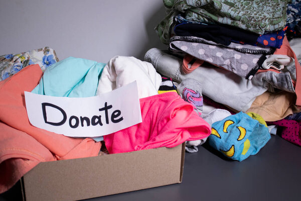 Donate sign handwritten with black letters. A box with clothes and a pile of clothes nearby on a grey table.clothes donation concept. copy space