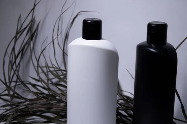 Samenstelling van twee witte en zwarte shampoo flessen zonder label in de buurt van gedroogde bladeren. lichaamsverzorging en schoonheidsconcept — Stockfoto