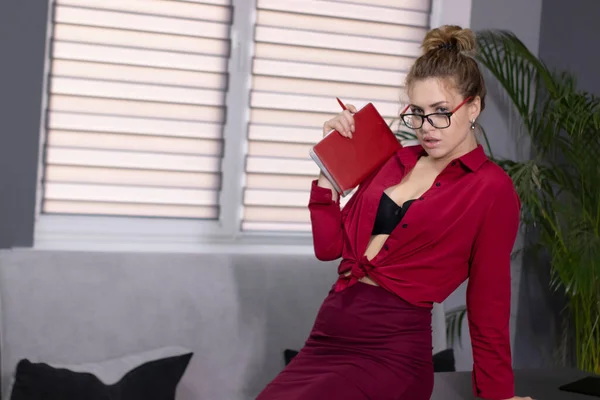 Düğmesiz kırmızı gömlekli, bordo etekli ve elinde kırmızı defter olan seksi bir sarışın. Gri ofiste