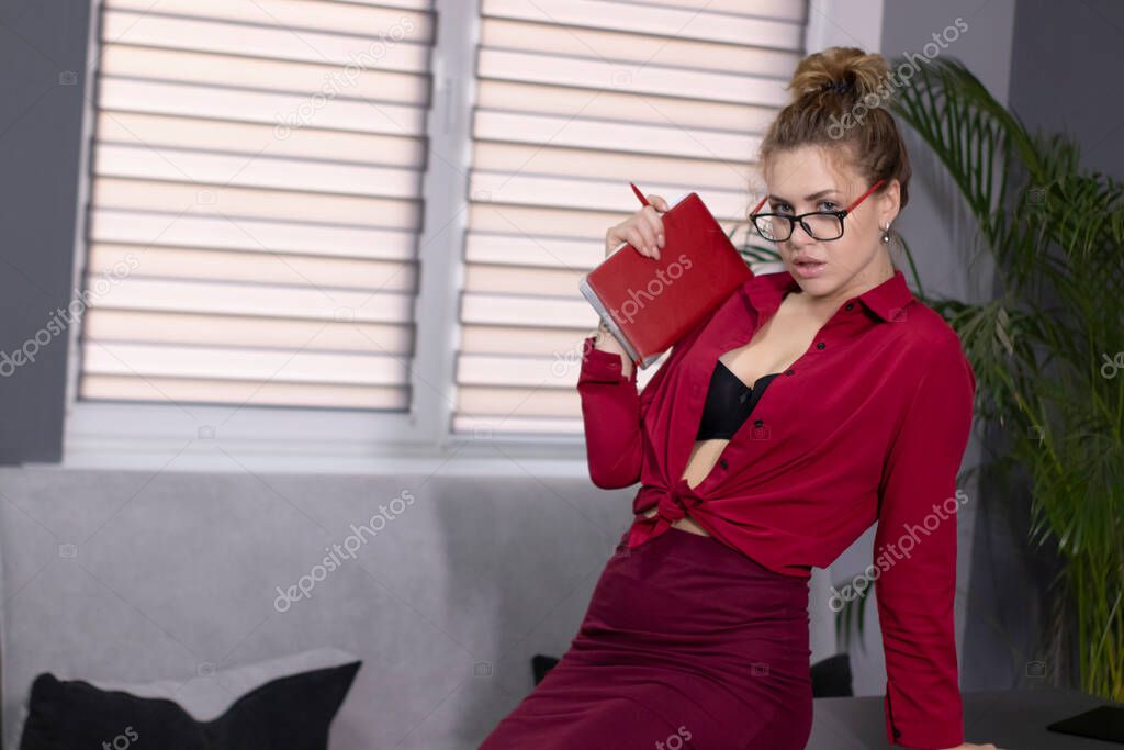 Sexy blonde in an unbuttoned red shirt, burgundy skirt and red glasses with a red notebook in her hands. In gray office