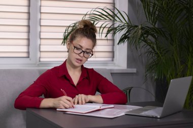 Kırmızı gömlekli ve kırmızı gözlüklü sarışın ofiste ya da evde çalışıyor. Uzak çalışma ya da öğrenim. Gri bir dizüstü bilgisayarla