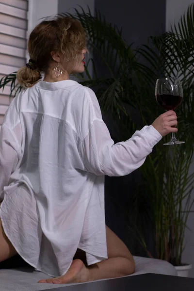 Sexy, bella ragazza bionda in un body nero, una camicia bianca e occhiali da sole con un bicchiere di vino rosso su un divano grigio. — Foto Stock