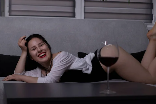Sexy, attraktive Brünette im schwarzen Body und weißem Hemd mit einem Glas Rotwein auf einem grauen Sofa — Stockfoto