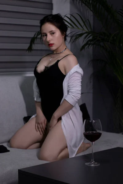 Sexy, jolie fille brune en body noir et une chemise blanche avec un verre de vin rouge sur un canapé gris — Photo