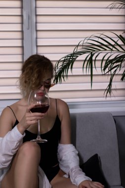 Siyah iç çamaşırlı, beyaz gömlekli ve güneş gözlüklü, gri kanepede bir kadeh kırmızı şarap içen seksi, güzel sarışın kız.