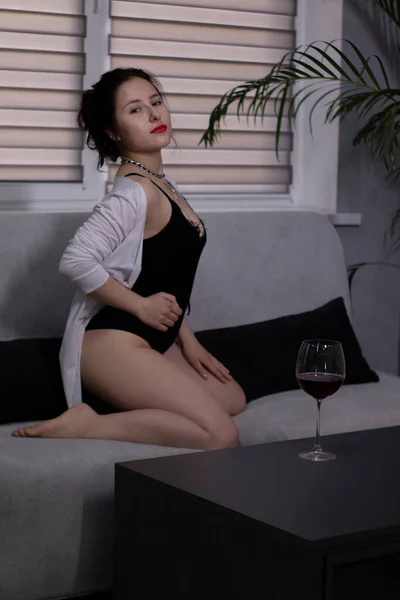 Sexy, attractive brunette girl in a black bodysuit and a white shirt with a glass of red wine on a gray sofa — Stock Photo, Image