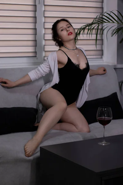 Menina morena sexy e atraente em um bodysuit preto e uma camisa branca com um copo de vinho tinto em um sofá cinza — Fotografia de Stock