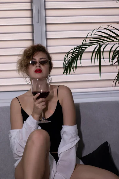 검은 보디 복을 입은 섹시하고 아름다운 금발의 여자, 흰 셔츠와 회색 소파에 빨간 와인 잔 이 있는 선글라스 — 스톡 사진