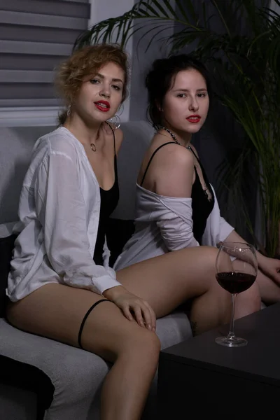 Dos chicas rubias y morenas- en lencería negra y una camisa blanca con copas de vino tinto en sofa.Novias, amantes —  Fotos de Stock