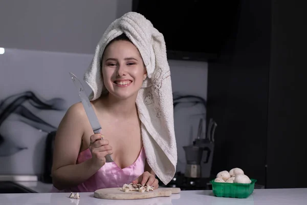 Привлекательная брюнетка - в розовой пижаме и с белым полотенцем на голове - резка грибов - на серой кухне . — стоковое фото