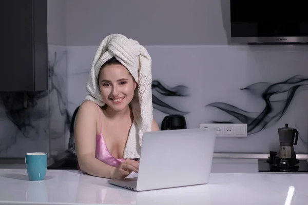 Fille brune - en pyjama rose et avec une serviette blanche sur la tête - avec tasse bleue, travaille sur un ordinateur portable télécommandé travail ou étude — Photo