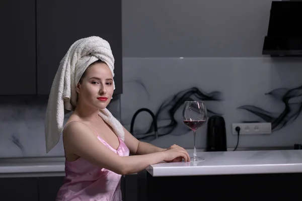 Jolie fille brune en pyjama rose et avec une serviette blanche sur la tête avec un verre de vin rouge dans la cuisine — Photo