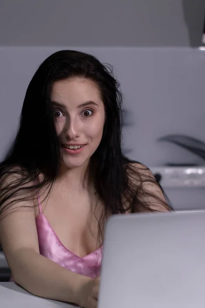 Menina morena em pijama rosa com uma xícara de chá-café azul, trabalha em um laptop - trabalho remoto ou estudo - em uma cozinha cinza — Fotografia de Stock