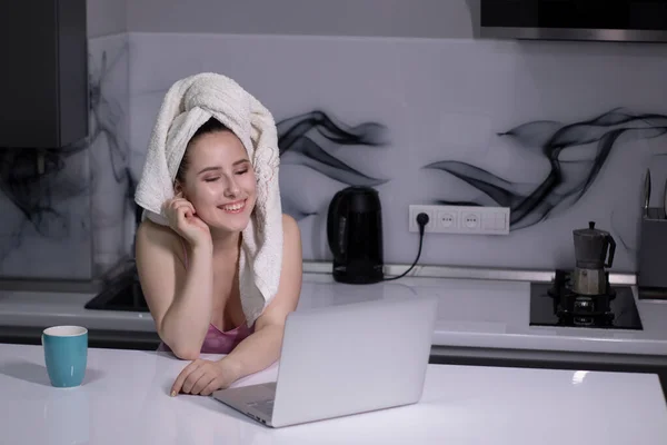 Ragazza bruna - in pigiama rosa e con un asciugamano bianco sulla testa - con coppa blu, lavora su un lavoro o uno studio a distanza per computer portatile — Foto Stock
