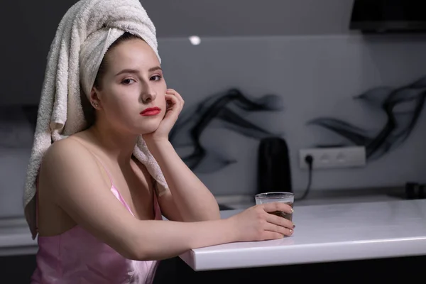 Atractiva chica morena - en pijama rosa y con toalla blanca en la cabeza - con vaso de whisky, en la cocina —  Fotos de Stock