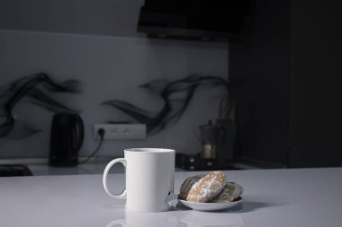 Beyaz bardakta çay ve çay poşeti. Gri mutfakta, beyaz masada zencefilli ekmeğin yanında.
