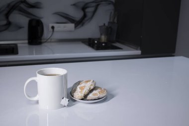 Beyaz bardakta çay ve çay poşeti. Gri mutfakta, beyaz masada zencefilli ekmeğin yanında.