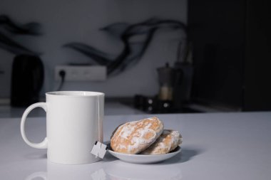 Beyaz bardakta çay ve çay poşeti. Gri mutfakta, beyaz masada zencefilli ekmeğin yanında.