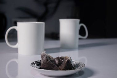 Gri mutfaktaki beyaz masada kullanılmış çay poşetlerinin yanında iki beyaz çay fincanı.