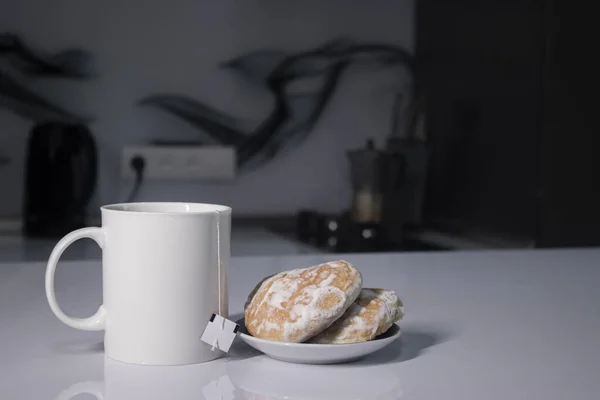 Bílý šálek s čajem a vystrkující čajový sáček vedle perníku na bílém stole v šedé kuchyni — Stock fotografie
