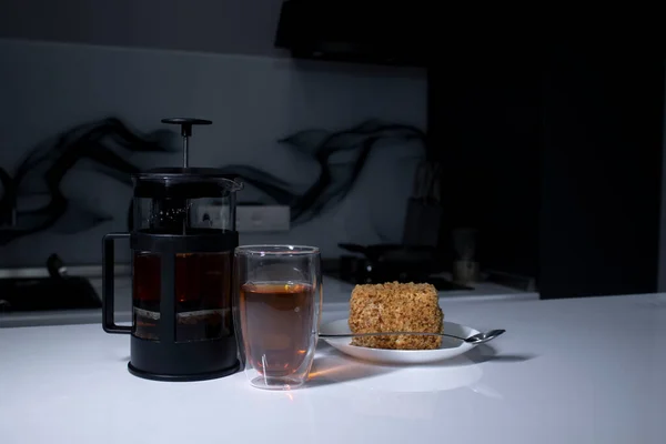 Šálek čaje v double-botoom sklenice, francouzsko-lis konvici a kousek dortu na bílém stole v šedé kuchyni — Stock fotografie