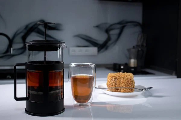 Cup of tea in a double-botoom glass, french-press teapot and piece of cake on white table in gray kitchen — Stock Photo, Image