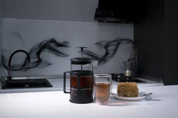 Kopje thee in een dubbel-botoom glas, franse-pers theepot en een stuk taart op witte tafel in grijze keuken — Stockfoto