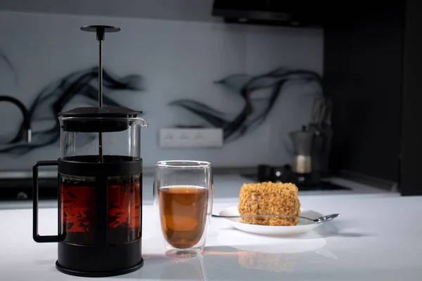 Cup of tea in a double-botoom glass, french-press teapot and piece of cake on white table in gray kitchen — Stock Photo, Image