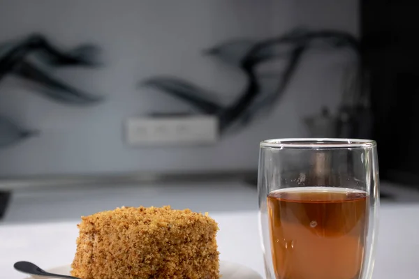 Cup of tea in a double-botoom glass and piece of cake on white table in gray kitchen — Stock Photo, Image