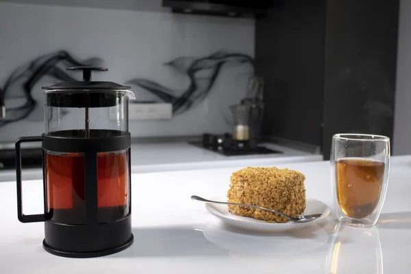 Kopje thee in een dubbel-botoom glas, franse-pers theepot en een stuk taart op witte tafel in grijze keuken — Stockfoto