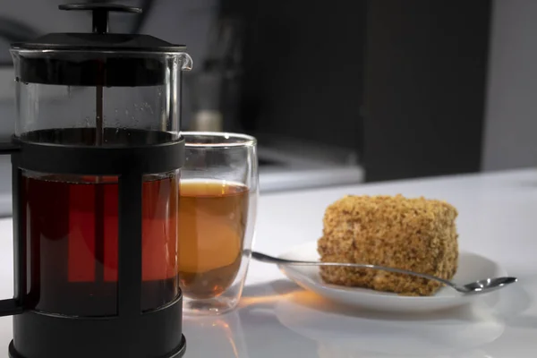 Cup of tea in a double-botoom glass, french-press teapot and piece of cake on white table in gray kitchen — Stock Photo, Image